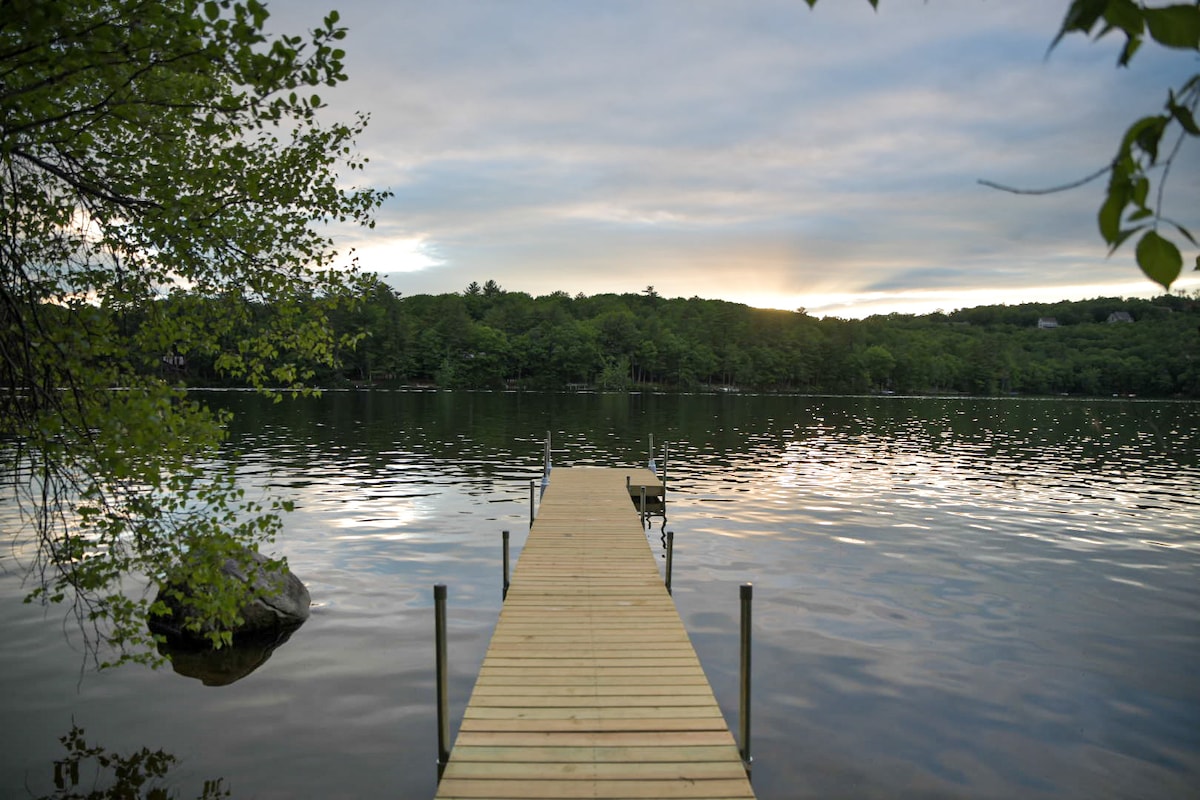 A wooden dock extends into the calm waters of Crystal Lake, framed by lush green trees on the opposite shore. The sun is setting behind the hills, casting gentle reflections on the water's surface, creating a serene atmosphere.