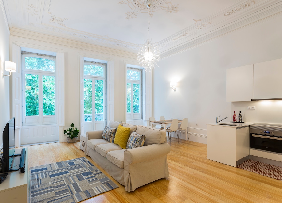 A spacious living area features large windows that allow natural light to brighten the room. A comfortable sofa is placed in front of a TV, while a dining table for four is visible nearby. The kitchenette is equipped with modern appliances against a clean backdrop.