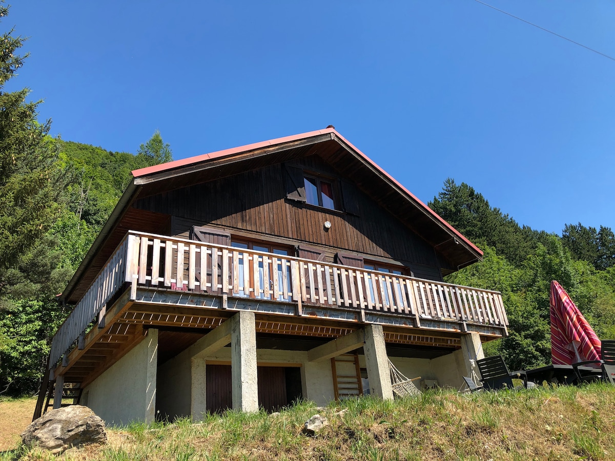 A two-story wooden chalet is featured, surrounded by lush greenery. A spacious balcony runs along the front, offering views of the natural landscape. The structure is positioned on a gentle slope, with a clear blue sky above and trees framing the scene.