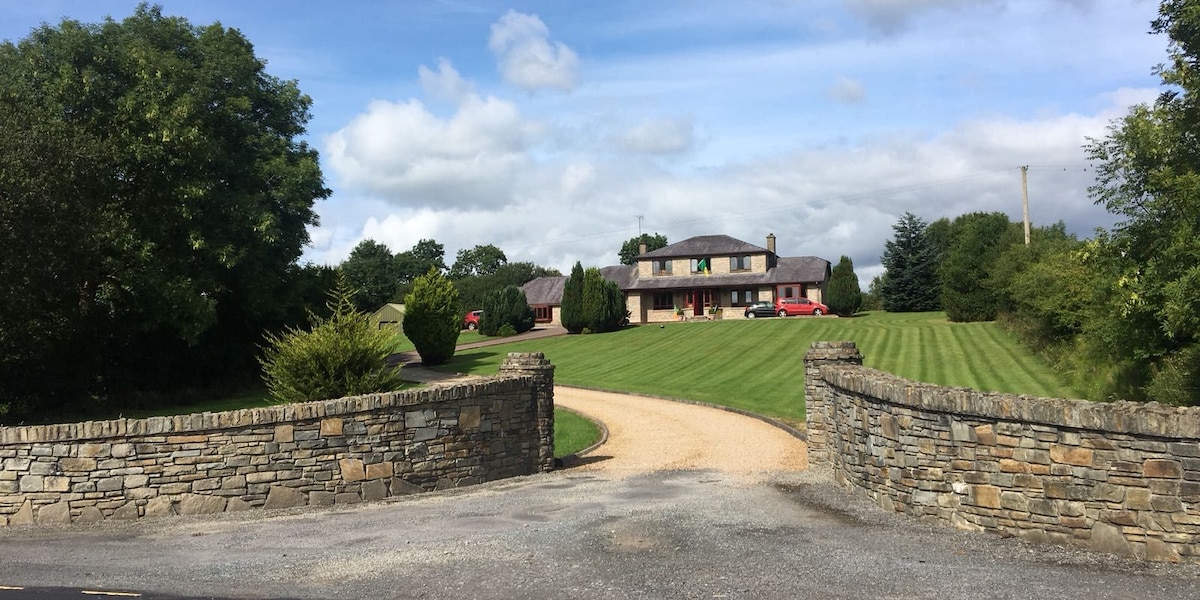 A panoramic view of a spacious property is captured, showing a well-maintained lawn and trees surrounding the house. The entrance is marked by a stone wall and a gravel path leading up to the two-story residence, which features a clean exterior and ample windows.