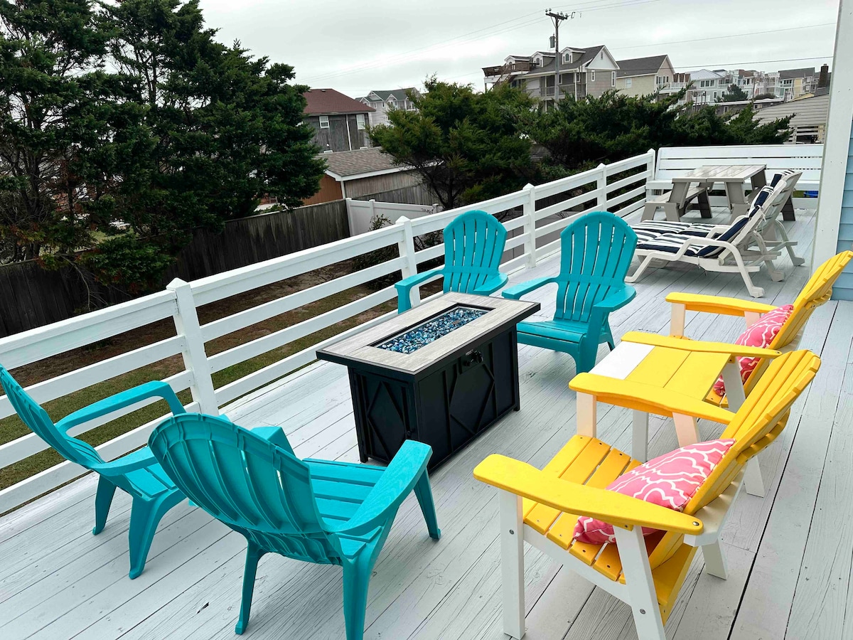 A spacious deck features a mix of brightly colored lounge chairs in turquoise and yellow, surrounding a fire pit with a glass top. A table with chairs is positioned in the background, with lush green trees visible along the boundary.