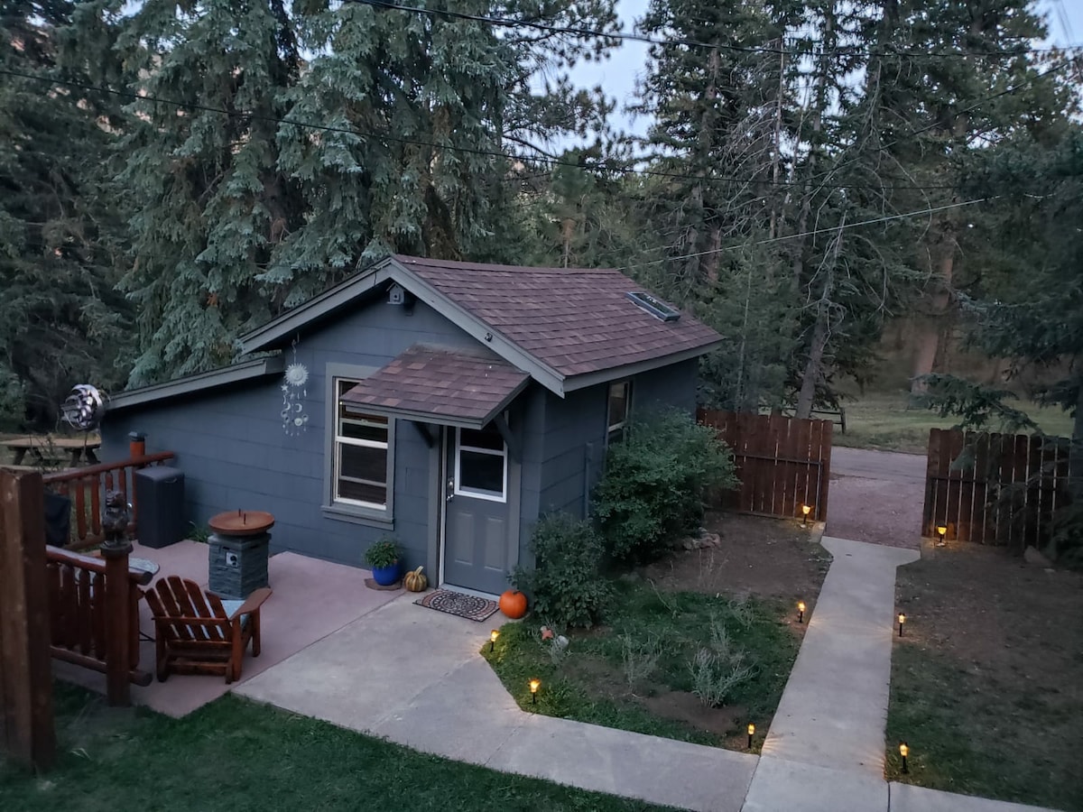 A charming cabin is nestled among tall pine trees, featuring a welcoming front porch adorned with seating. A stone path leads to a wooden fence, illuminated by small lights that enhance the cabin's tranquil setting. The exterior is painted in muted tones, blending with the natural surroundings.