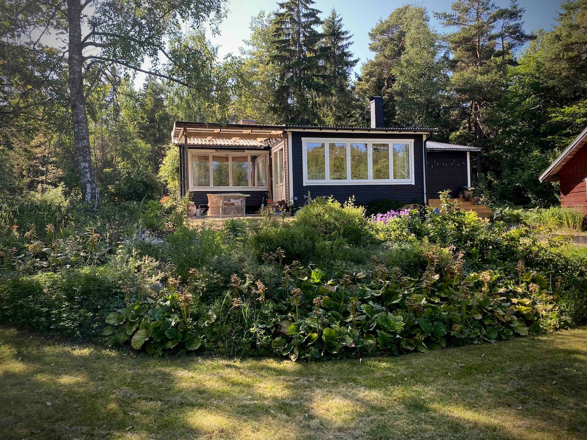 A charming cabin is nestled among lush greenery, surrounded by tall trees. Large windows allow natural light to fill the interior. A deck extends from the front, providing space for outdoor seating. Vibrant flower beds and foliage enhance the natural setting of the property.