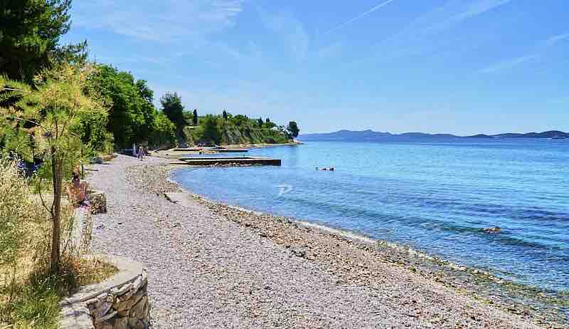 A serene beach scene features a pebbly shoreline beside calm, clear waters. Lush greenery lines the edge, providing a natural border. A few individuals can be seen enjoying the beach in the distance, while distant landforms create a peaceful backdrop under a clear blue sky.