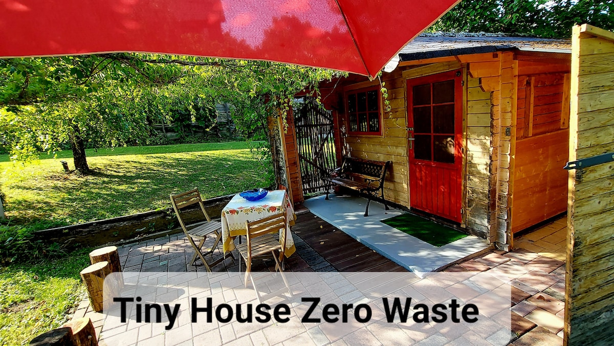 A wooden tiny house is set in a green outdoor space, featuring a red door and multiple windows. A shaded patio area is equipped with a table and chairs, while a small decorative rug adds color. Surrounding trees provide a natural backdrop.