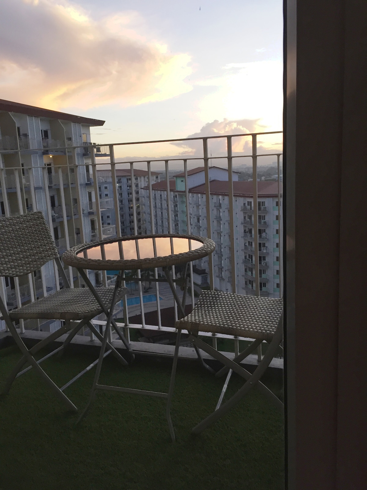 A small terrace is visible, furnished with two metal chairs and a round table. The scene captures a view of neighboring buildings and a distant glimpse of the sunset sky. The terrace has a green carpet, adding a touch of comfort to the outdoor space.