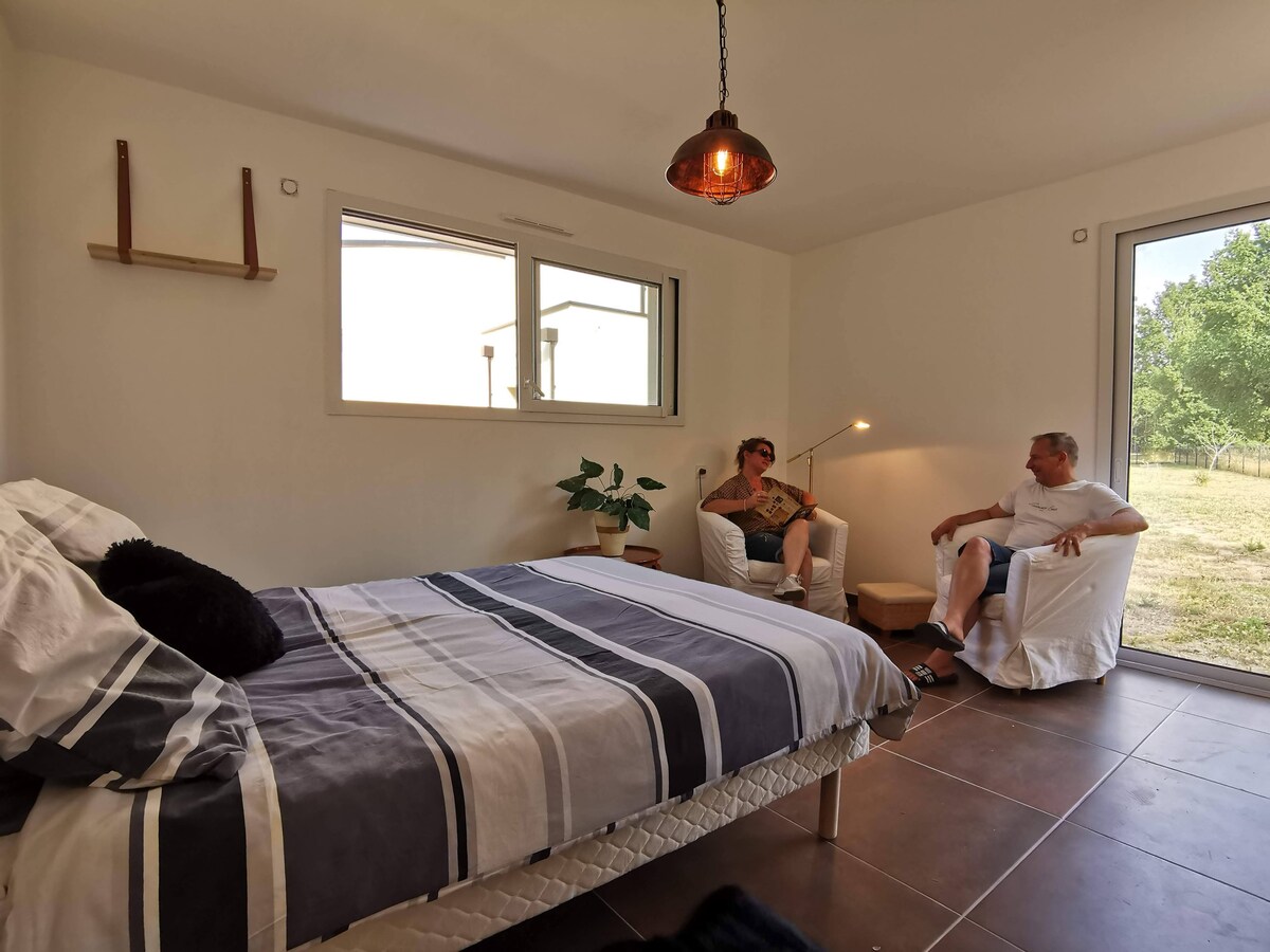 A spacious room features a comfortable bed with striped bedding and a cozy armchair set beside a standing lamp. Bright light enters through large windows, offering views of the surrounding greenery. A small plant is positioned near the bed, adding a touch of nature.