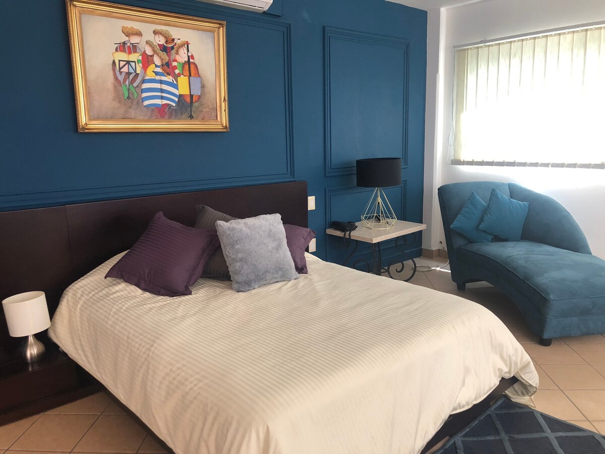 A spacious bedroom is showcased with a large bed dressed in white linens and assorted purple pillows. A stylish blue chaise lounge sits nearby, accompanied by a small side table. Natural light enters through a window adorned with vertical blinds, highlighting the textured blue wall and an art piece above the bed.