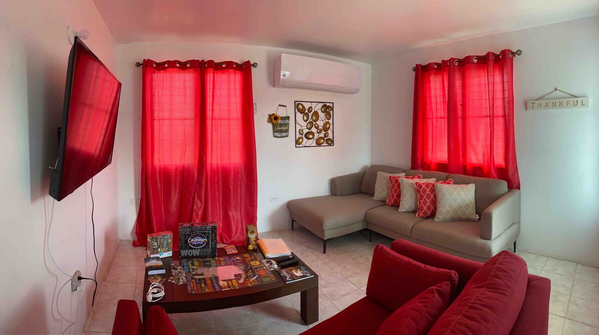 A cozy living area is highlighted by bright red curtains and comfortable seating arrangements. A modern television is mounted on the wall, with a coffee table displaying various magazines and decorative items. Light fills the space, enhancing the welcoming environment.