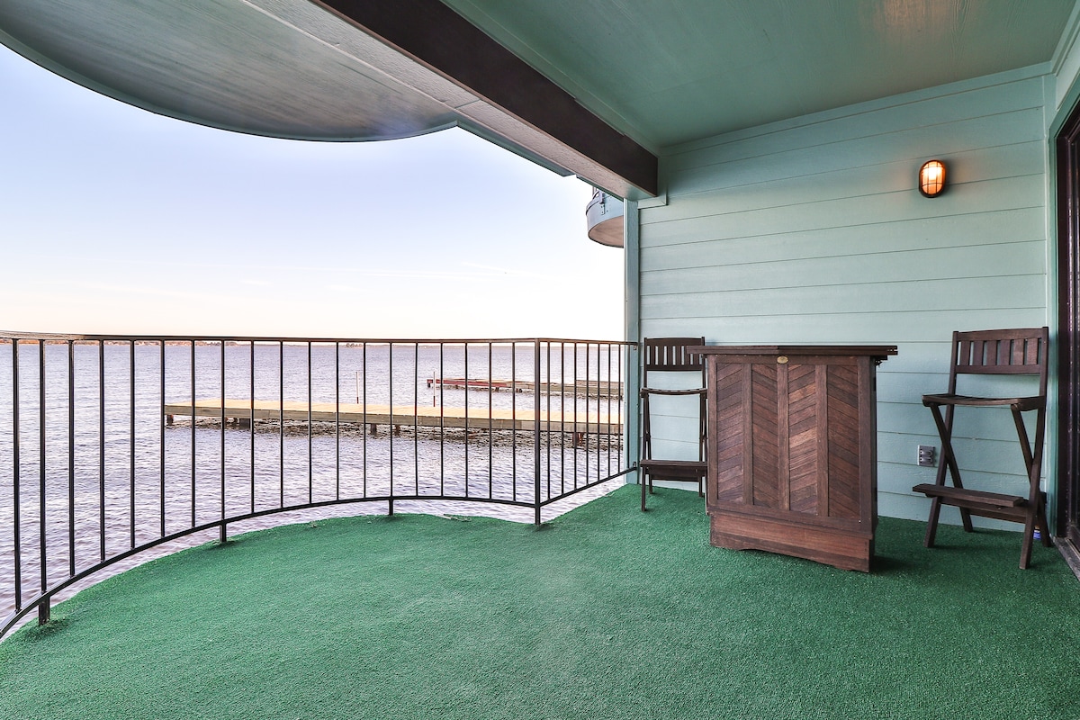 A private over-the-water balcony features a wooden table and two chairs, surrounded by a railing. The space is covered with green outdoor carpeting, offering a clear view of the lake and wooden pier in the distance, under a clear sky.