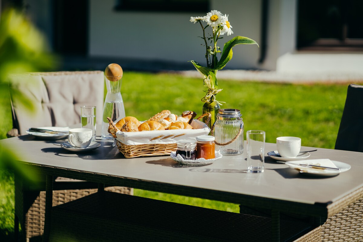 A well-set outdoor dining table features a basket of fresh bread rolls, alongside a vase with a flower. Glassware and fine dishware are arranged neatly, with a glass carafe and jars containing spreads placed on the table. Lush green grass is visible in the background.