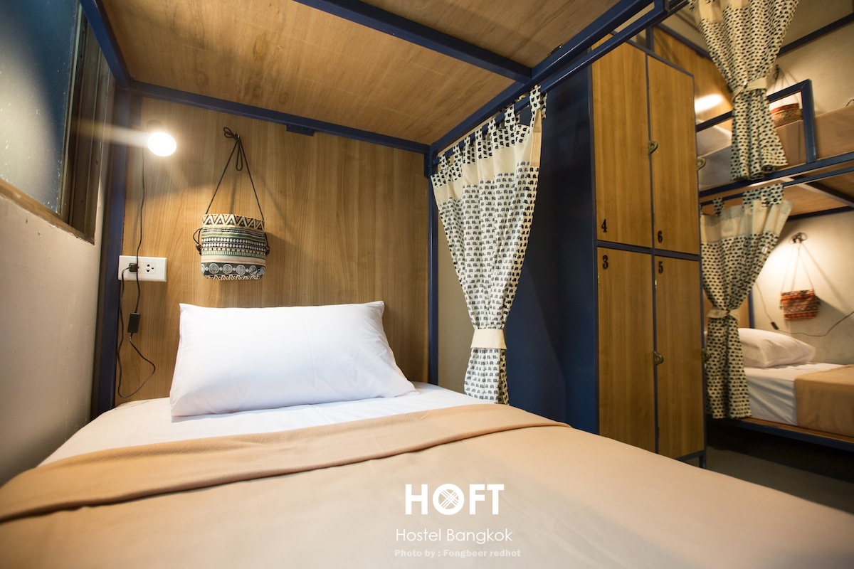 A cozy sleeping area features a soft bed dressed in neutral linens and positioned against a wooden wall. A decorative light fixture hangs overhead, while adjacent lockers provide secure storage for personal items. Nearby, a curtain partially separates the space from another sleeping area.