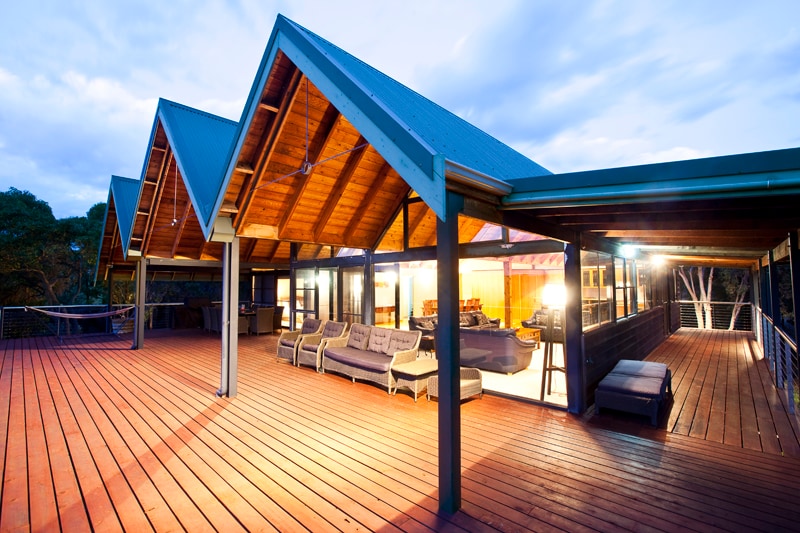 A spacious deck area features a wooden floor and an expansive roof with pitched peaks. Comfortable lounge seating is arranged on one side, while the warm glow from interior lighting invites relaxation. Surrounding trees create a natural backdrop, enhancing the serene atmosphere of the space.