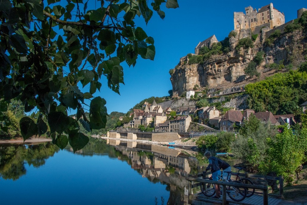 A serene view is captured along the river, reflecting the historic village and its stone buildings. A castle is perched atop a cliff, with lush greenery surrounding the area. A person is seen near a wooden dock with bicycles, adding to the tranquil scenery.