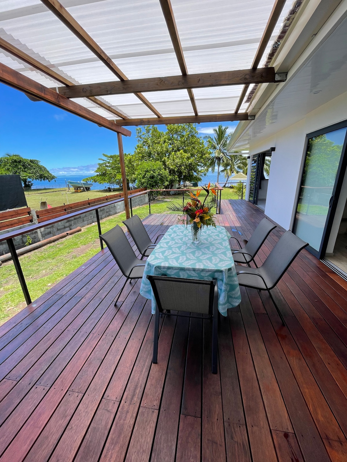 A spacious wooden terrace features a dining table set for six, adorned with a colorful centerpiece. The area is shaded and offers a view of the surrounding greenery and sea through large glass doors, providing an inviting space for outdoor dining.