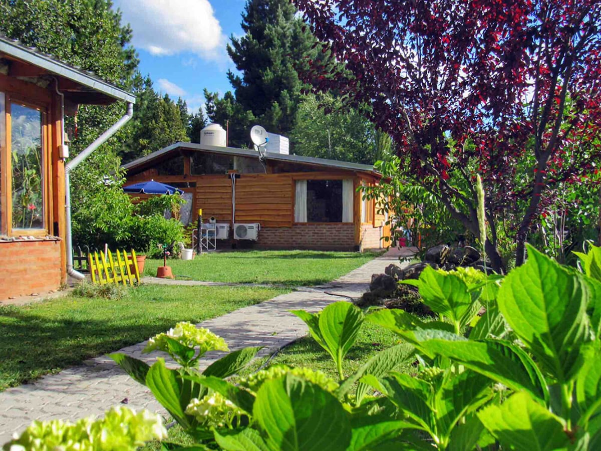 A bright, independent cabin is surrounded by well-kept gardens and lush greenery. A stone pathway leads to the cabin, with various plants and colorful foliage visible. The cabin features large windows and outdoor seating, contributing to a serene outdoor environment.