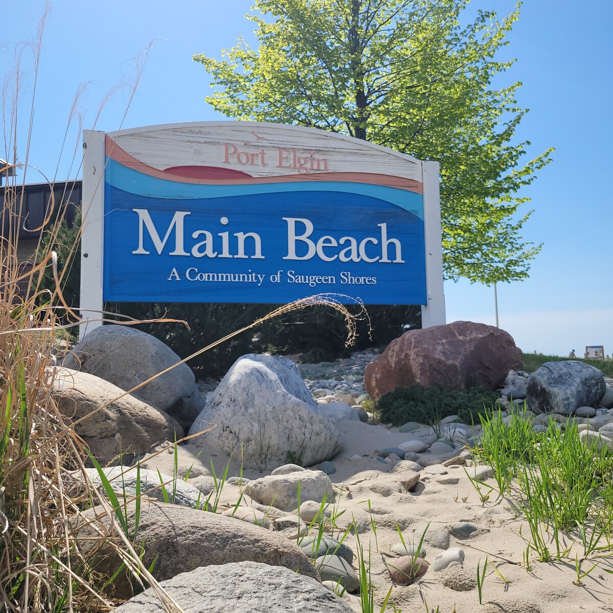 A welcoming sign for Port Elgin Main Beach is prominently displayed, surrounded by natural elements such as smooth stones and grassy patches. Bright blue paint highlights the beach's name, while a clear sky and lush trees are partially visible in the background.