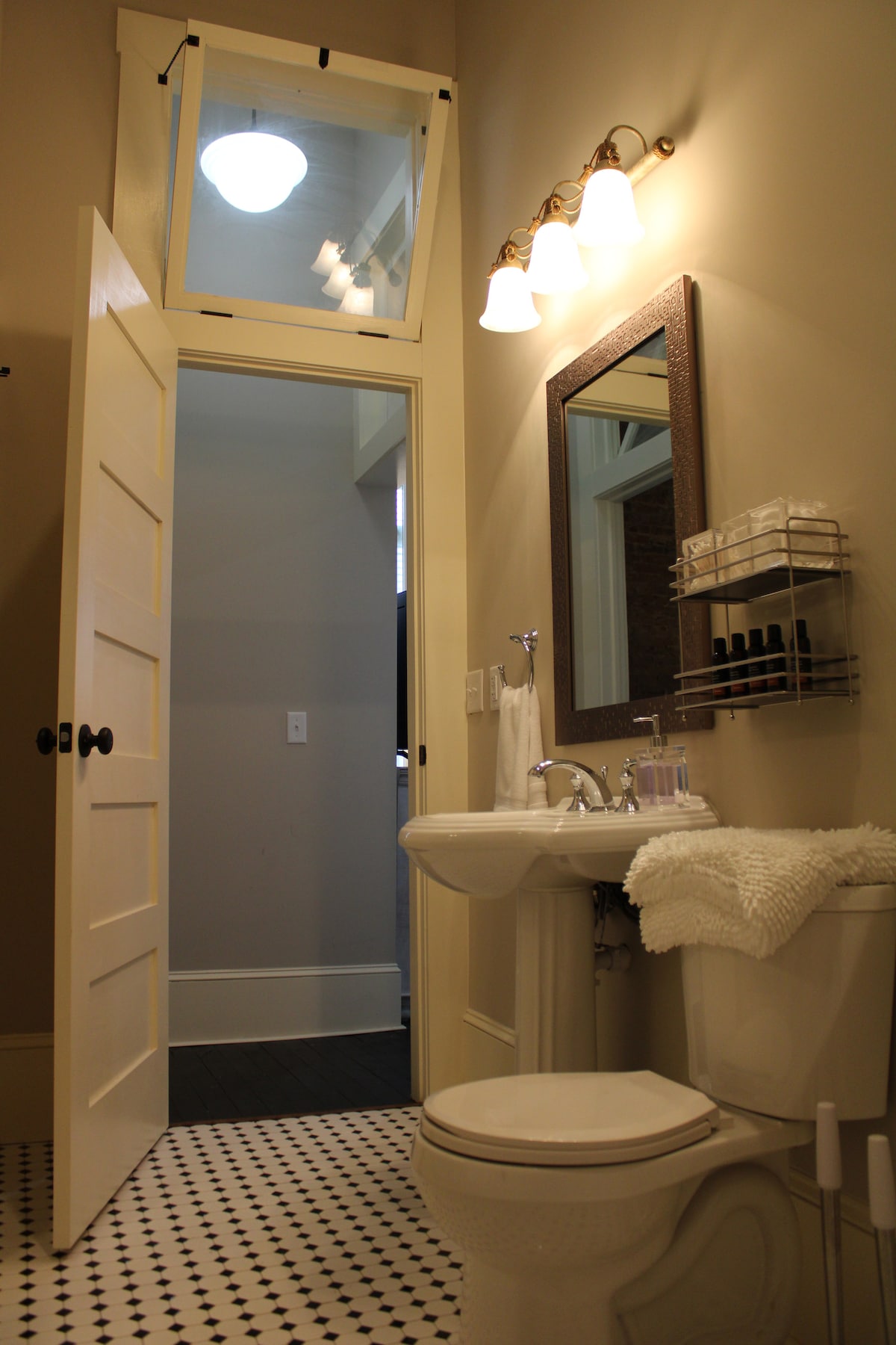 A well-lit bathroom is featured, showcasing a white porcelain sink and toilet. A mirror hangs above the sink, complemented by a decorative shelf with toiletries. Black and white tile flooring adds a classic touch, while a door leads to the adjoining room.