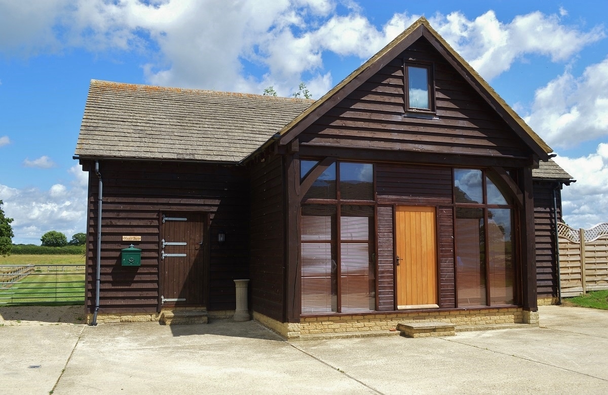 The exterior of The Manor Barn is captured, showcasing a wooden structure with a steeply pitched roof and large windows. A centrally located wooden door is framed by a stone porch. The surroundings feature well-maintained grassy fields under a partly cloudy sky.