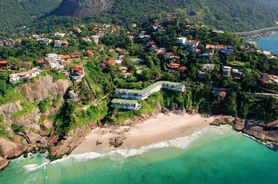 An aerial view captures the coastal landscape with lush greenery and residential homes nestled among rocky hills. The soft sandy beach stretches along the turquoise waters of the ocean, creating a serene contrast against the vibrant foliage and cliffs of the surrounding area.