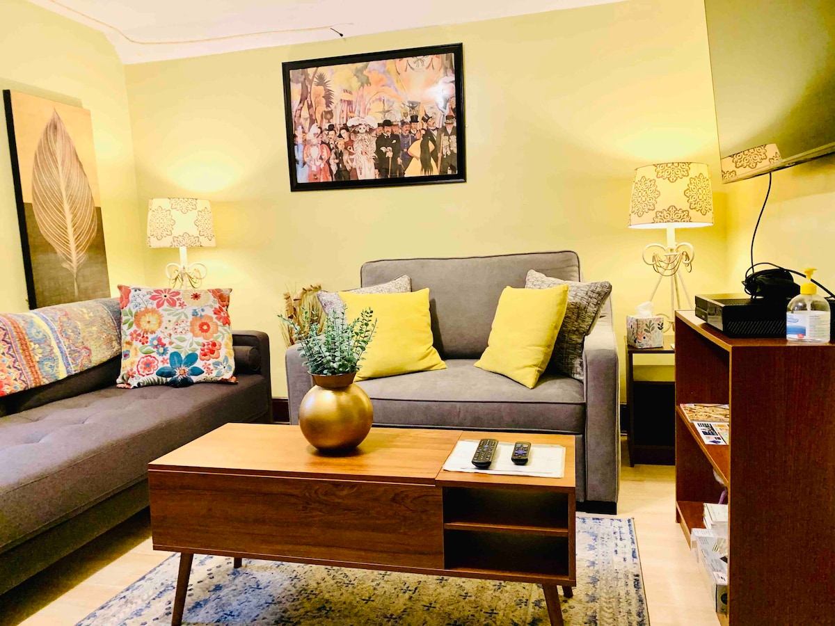 A living area features a gray sofa with colorful patterned cushions and a matching armchair. A wooden coffee table with decorative items is positioned between the seating. A framed artwork adorns the wall, and soft lighting is provided by two stylish lamps.