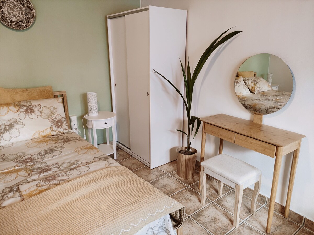 A bright sleeping area features a spacious bed with floral bedding, complemented by a light-colored side table. A wooden desk with a round mirror and a potted plant adds to the decor. A white wardrobe is positioned nearby, providing additional storage.