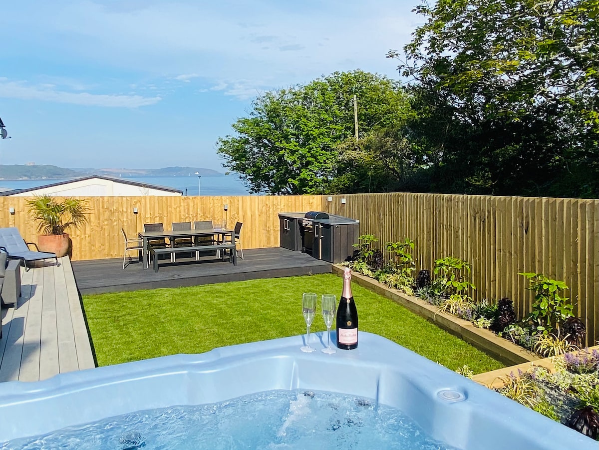 An outdoor space is showcased with a hot tub in the foreground, accompanied by a bottle of champagne and two glasses. A dining area with a table and chairs is visible on a raised deck, framed by lush greenery and a wooden fence. The serene view of water and hills is seen in the background.