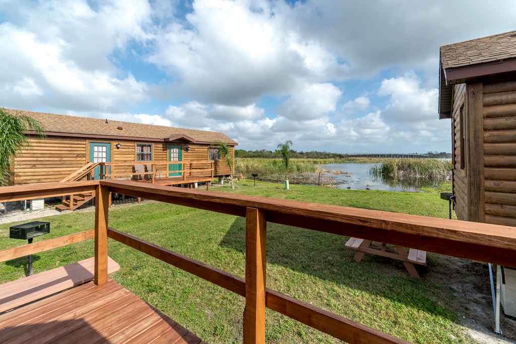 A spacious deck is seen in the foreground, featuring wooden railings that provide a view of the lake and grassy area. Log cabins are visible in the background, with cloudy skies above and tall grasses by the water's edge.