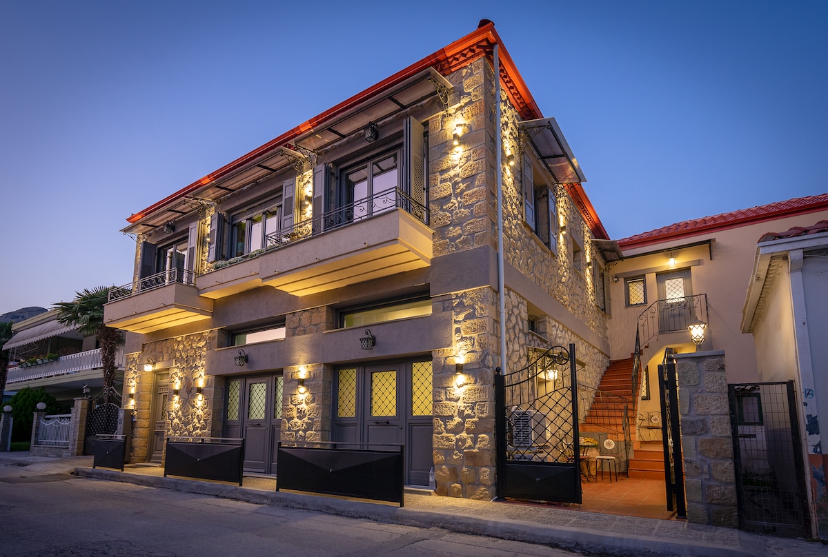 A two-story stone building is illuminated by warm lights at dusk. Balconies adorned with wrought iron railings offer an inviting aspect. The entrance features a staircase leading to upper-level access, while the surrounding area showcases a neat, paved pathway.
