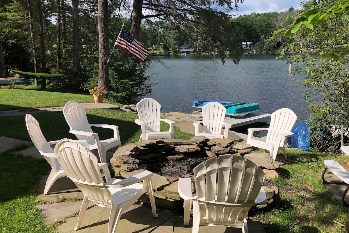 A fire pit surrounded by white Adirondack chairs is positioned by the lake. An American flag flutters nearby. The serene water reflects the greenery and sky, enhancing the tranquil setting. The area is well-maintained, with a path leading to the lake and lounging areas.