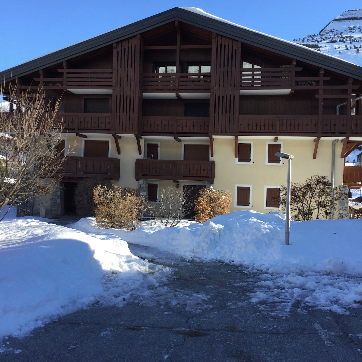 The residence features a three-story chalet-style building with a warm, wooden exterior. Snow blankets the front, and several balconies are visible, adorned with wooden railings. Surrounding pathways are cleared of snow, inviting access to the entrance.
