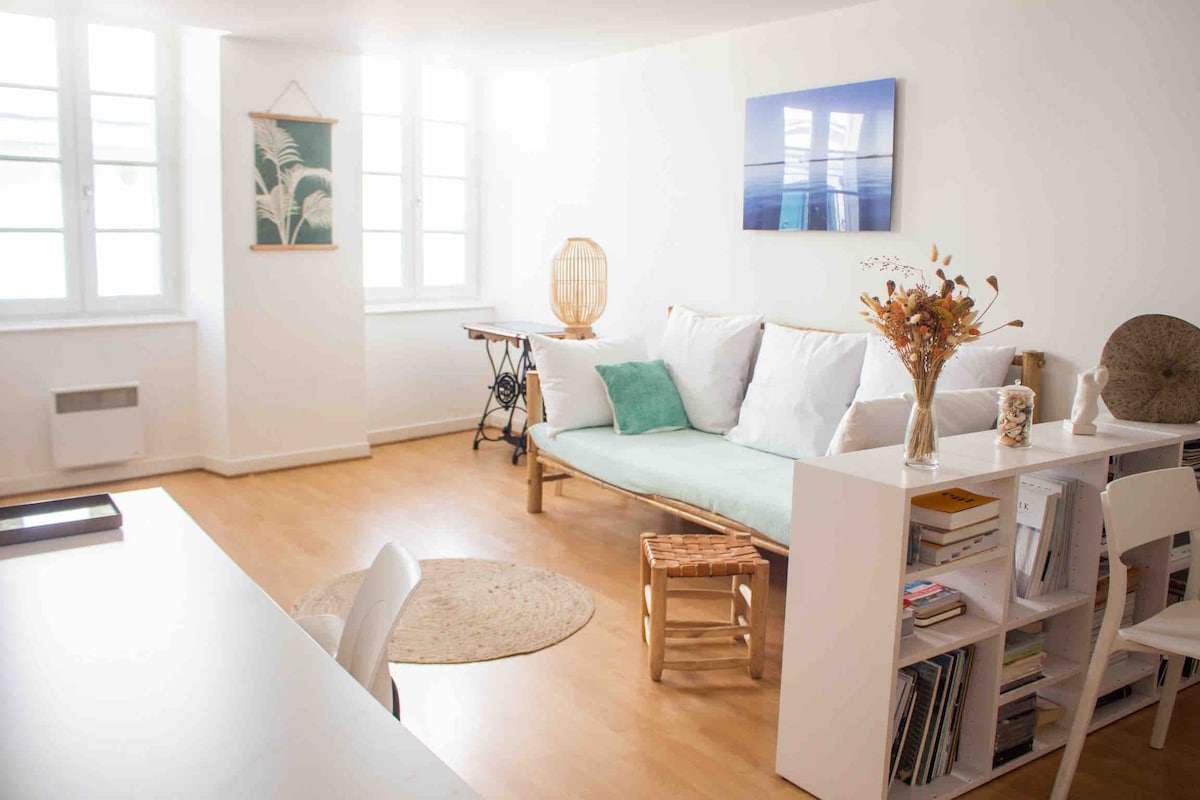 A light-filled living space features a comfortable sofa adorned with soft pillows, complemented by a side table and a rug. A small dining table is positioned near the window, and a bookshelf displays various books. Natural light filters through multiple windows, enhancing the airy atmosphere.