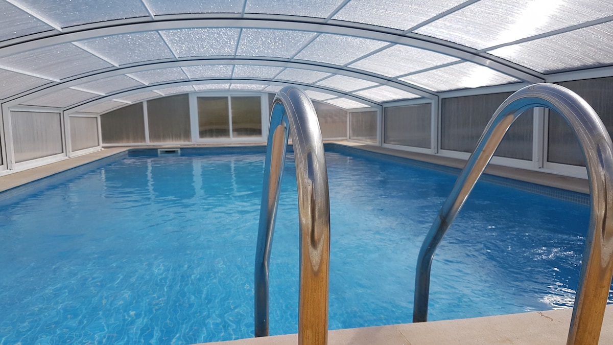 An indoor swimming pool is enclosed under a clear, arching cover. The pool features a smooth, blue surface, with a pair of metal handrails leading into the water. Light reflects gently off the water, creating a serene atmosphere.