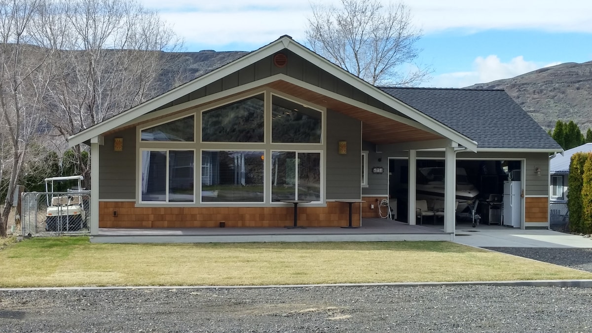 A modern single-story house is shown, featuring large front windows that allow natural light to illuminate the interior. The front porch is covered, providing shelter. A well-maintained lawn surrounds the property, with a gravel driveway leading up to the attached garage that accommodates a boat.