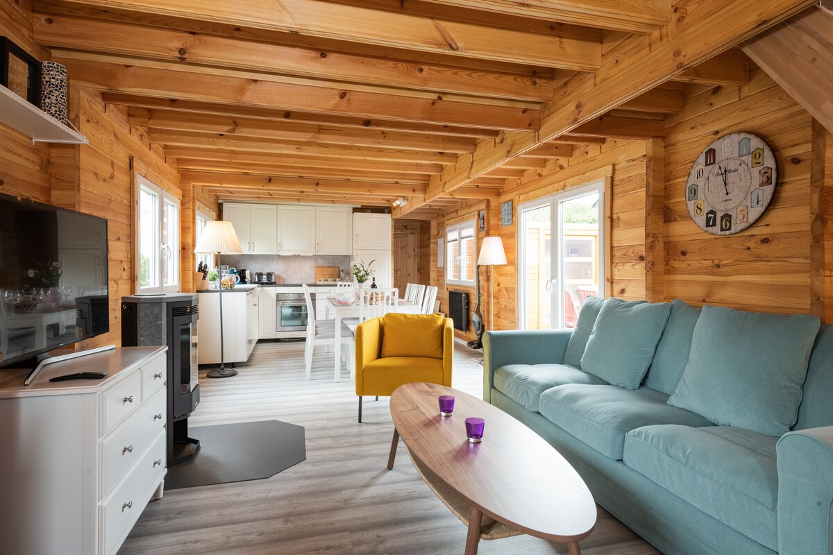 The open-plan living area features wooden walls and beams, enhancing the cozy atmosphere. A comfortable couch and a yellow armchair provide seating. A coffee table sits in front of the couch, with a modern flat-screen TV positioned nearby. Bright natural light fills the space through multiple windows.
