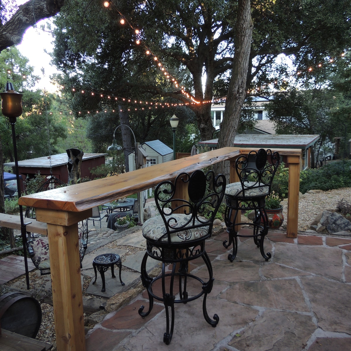 A custom cedar bar with a wooden countertop is surrounded by two wrought-iron chairs featuring cushions. Twinkling bistro lights hang overhead, illuminating the flagstone patio and creating a relaxed outdoor space. Natural rock and oak trees frame the view, enhancing the setting's tranquility.