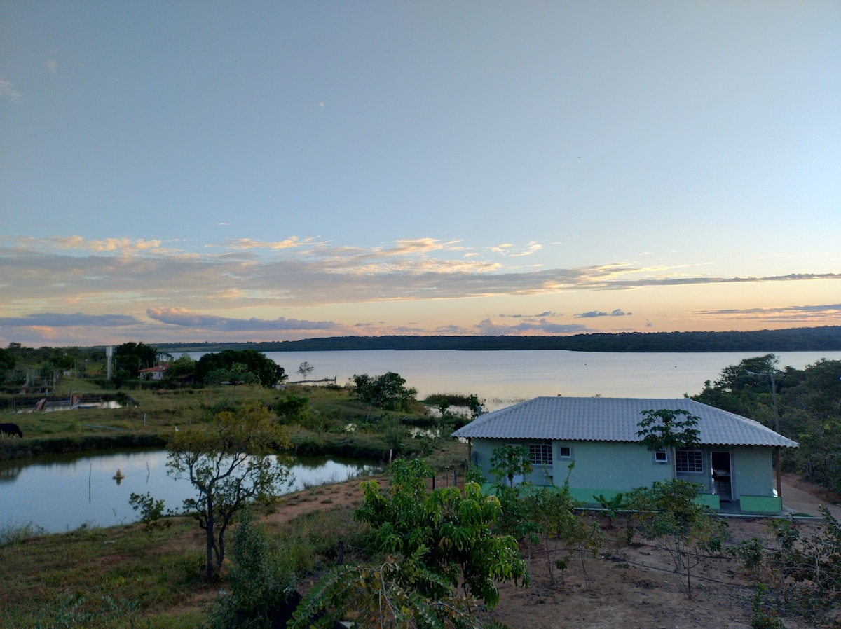A serene view of a tranquil lake is presented, with a charming green house situated nearby. Lush greenery is visible in the foreground, complementing the calm waters and soft sky hues at dusk.