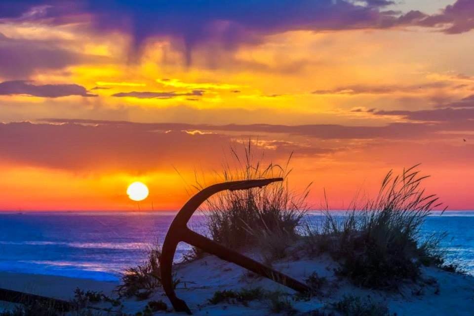 A scenic view of a vibrant sunset over the ocean showcases a warm blend of orange, pink, and purple hues. Silhouetted against the sky, an anchor is positioned in the foreground, partially surrounded by grass and sand.