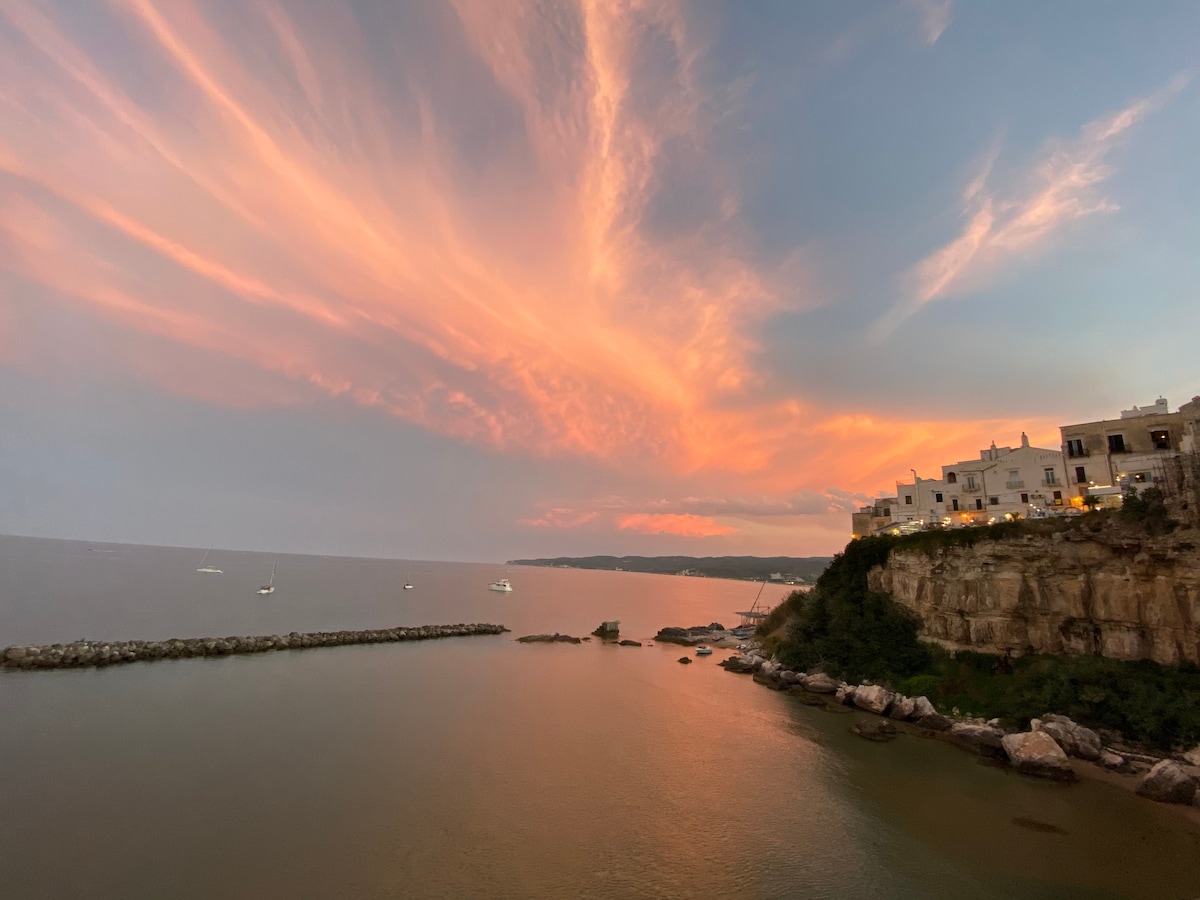 A sunset view over the tranquil sea shows a gradient of orange and pink hues in the sky. In the distance, boats can be seen on the water, while the silhouette of a coastal town is visible on the right, illuminated by soft lights.