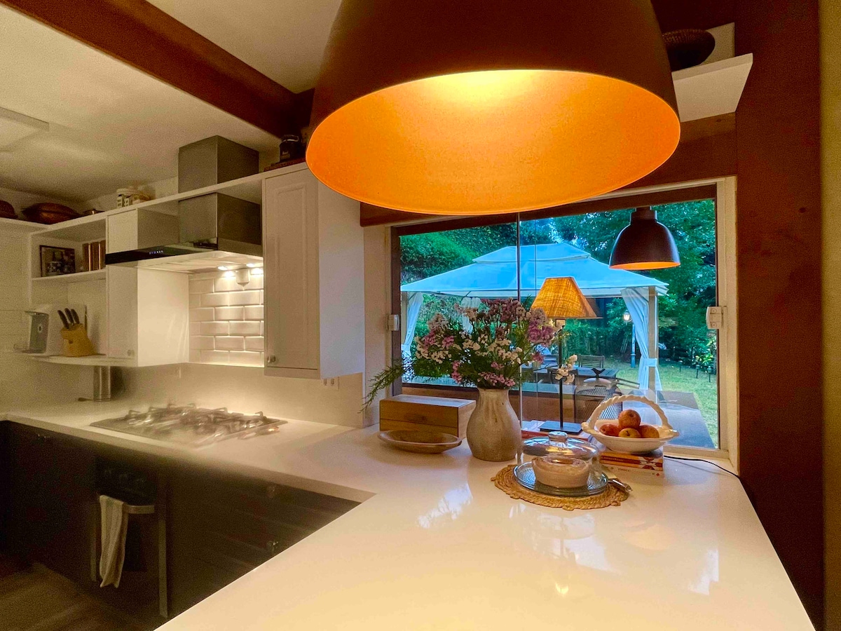 An inviting kitchen space is highlighted by bright, ambient lighting from a large pendant lamp. A window frames a view of an outdoor gazebo, while the countertop features a decorative arrangement of fresh flowers and fruits, suggesting a welcoming atmosphere for cooking and dining.
