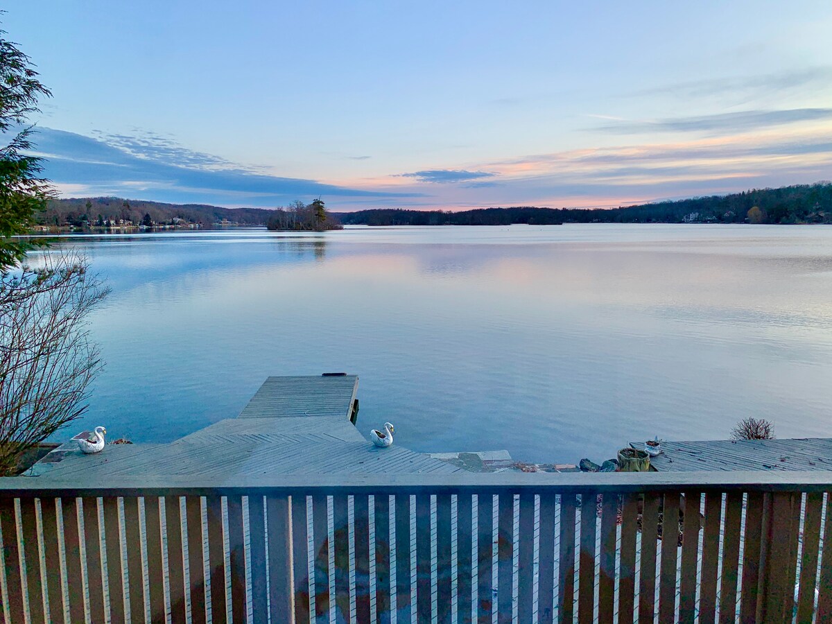 A serene lakeside view is captured from the deck, showcasing still waters reflecting the pastel colors of dawn. A wooden dock extends into the lake, with two swans gracefully positioned nearby, surrounded by gentle ripples and lush greenery.