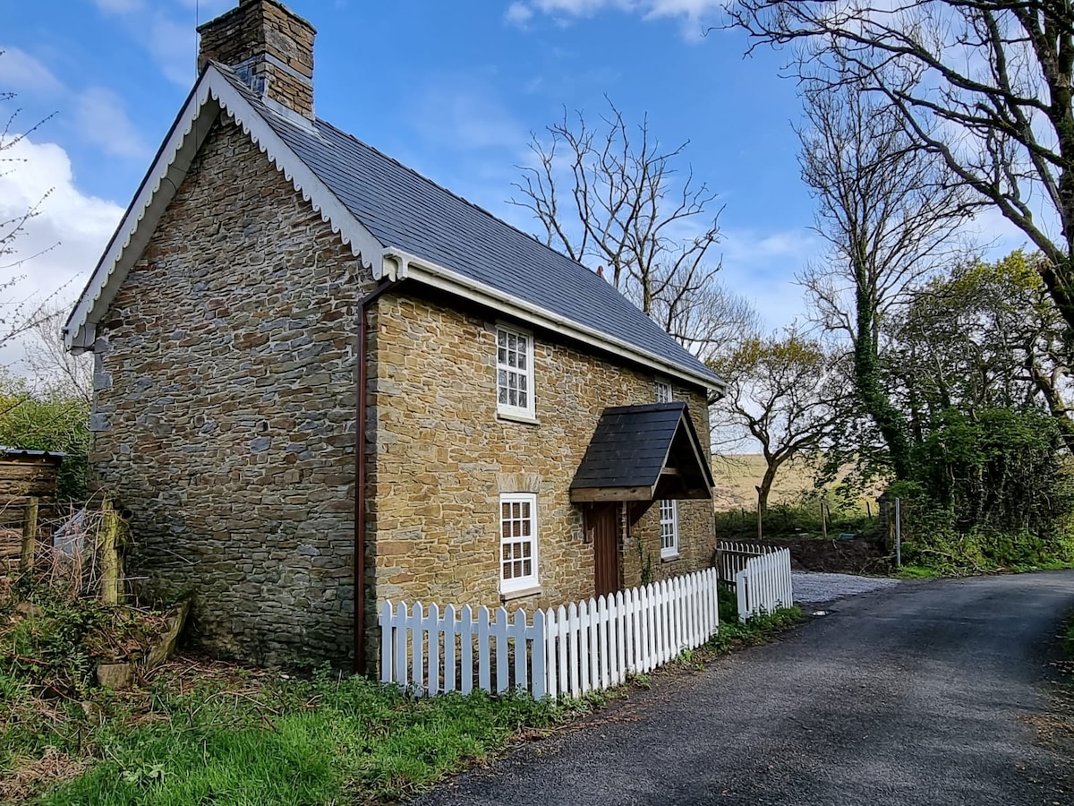 The exterior of a charming stone cottage is presented, featuring a pitched roof and white trim around the windows. A small, white picket fence surrounds the entrance, with a gravel path leading to the door. Lush greenery and trees are visible in the background.