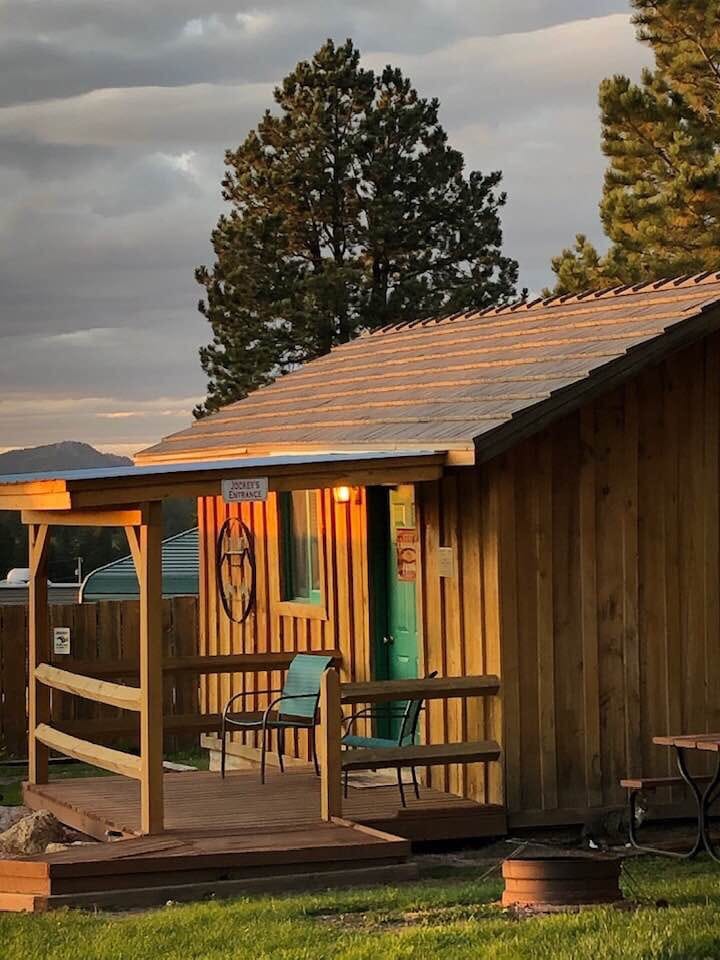 A wooden cabin features a small porch with two chairs and a decorative sign. The cabin is illuminated by warm light, highlighting the natural wood texture, while a fire ring sits nearby in the grassy area. Tall trees and distant hills are visible in the background.