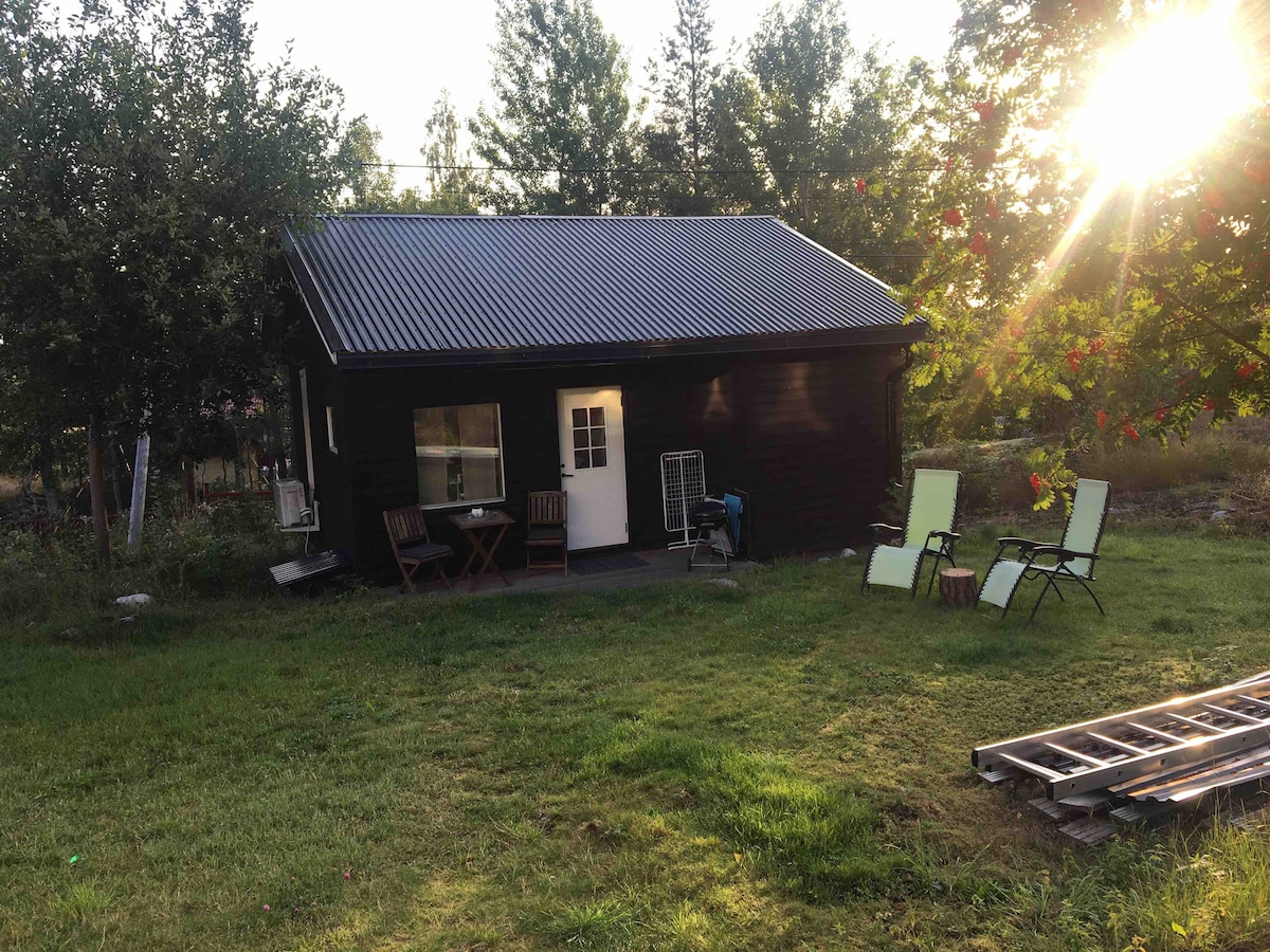 A small cabin with a dark wood exterior features a front door flanked by two chairs on a green lawn. A table with two chairs is set outside, and a light spills from the windows as the sun sets among the trees.