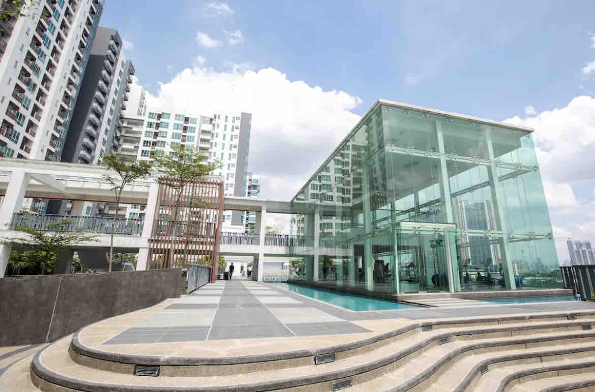 A modern building with extensive glass walls is situated beside a tranquil pool area. Steps lead from the paved pathway to the water's edge, surrounded by greenery. The surrounding high-rise structures are visible against a bright blue sky with fluffy clouds.