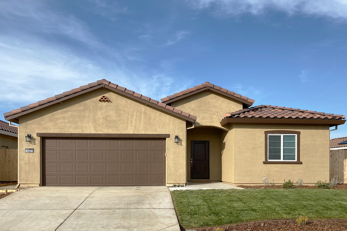 A single-story home is displayed, featuring a stucco exterior with a brown tiled roof. A driveway leads to a two-car garage, and a landscaped front yard is visible. Multiple windows allow natural light into the interior, and details such as decorative shutters enhance the entrance.