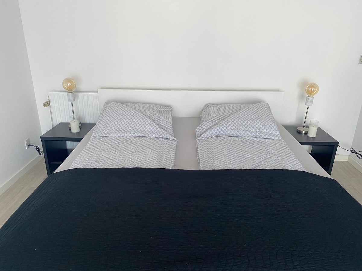 A spacious bed is positioned against a white wall, featuring two separate bedding setups. Nightstands are placed on either side, each with a table lamp and a candle. The bedding displays a subtle patterned design, complemented by a dark, textured blanket draped across the bottom.