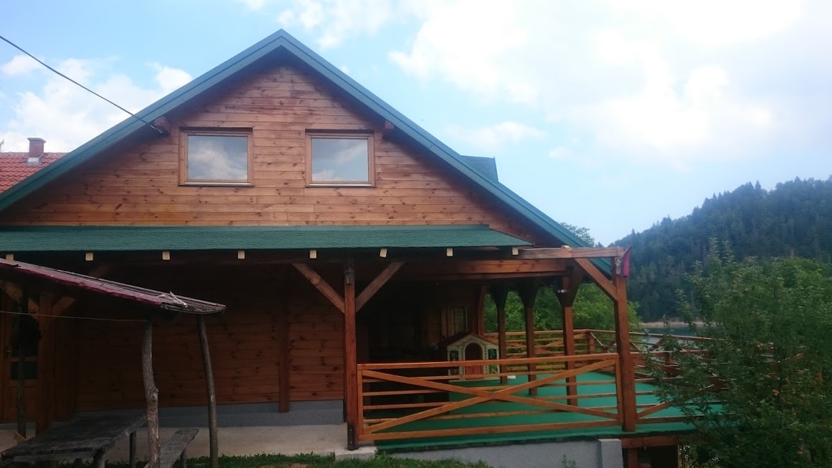 The wooden exterior of the house is highlighted, showcasing a sloped roof and two windows. A spacious porch with a railing provides access to the outdoors. Surrounding greenery and a glimpse of the lake enhance the natural setting.