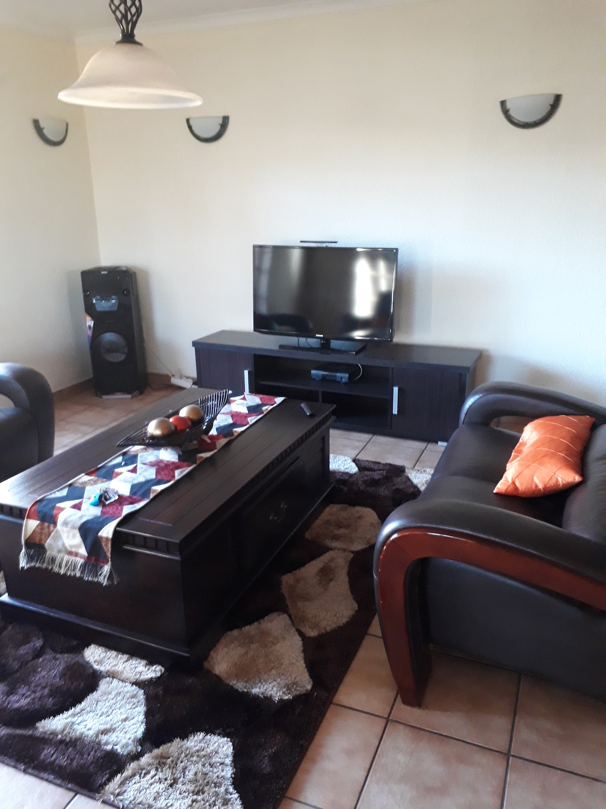 A living room features a dark wood coffee table at the center, adorned with decorative items. Two leather sofas provide seating, arranged around a patterned rug. A television rests on a black media console, with speakers positioned nearby. Soft light is provided by a hanging lamp.