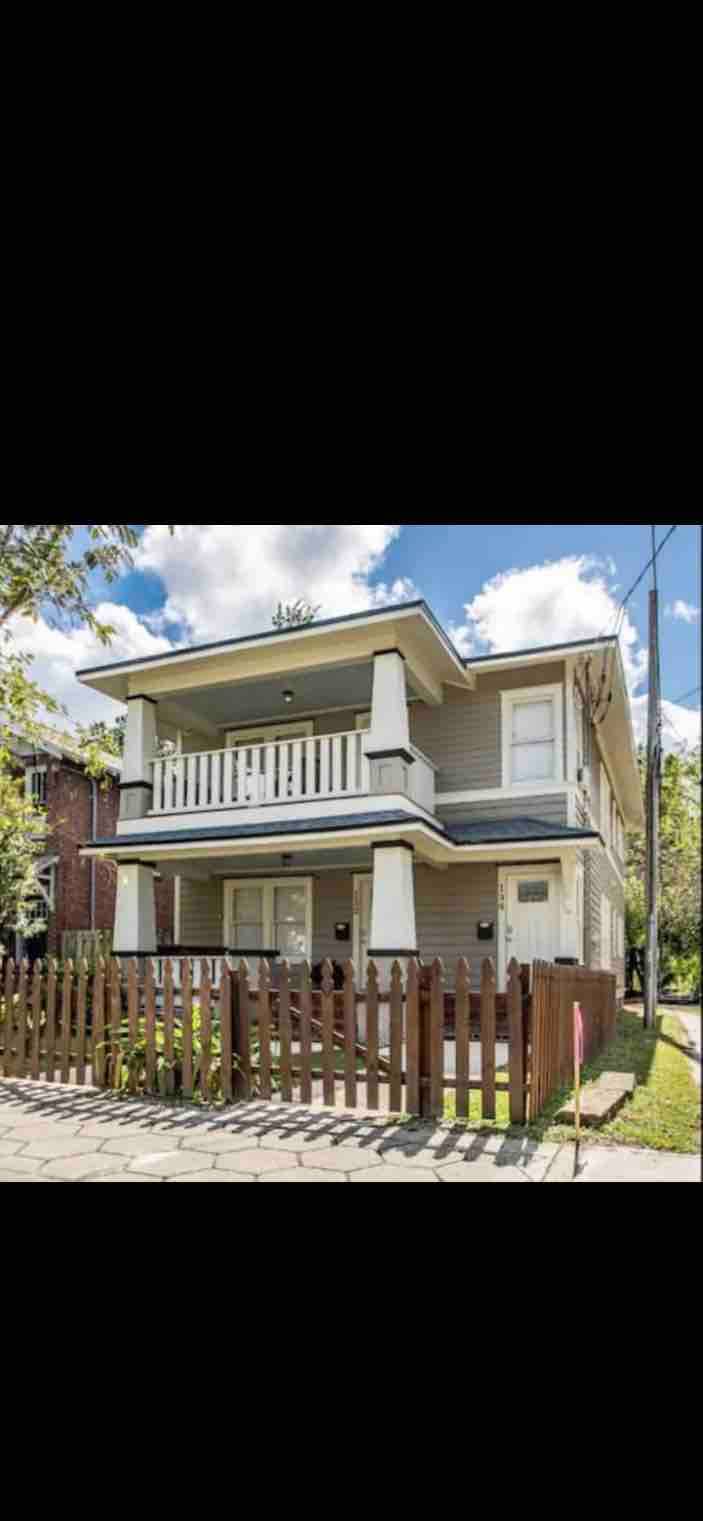 A two-story house features a light exterior with a covered porch and white columns. A wooden fence surrounds the front yard, with greenery visible. The sky above is partly cloudy, allowing natural light to illuminate the scene.
